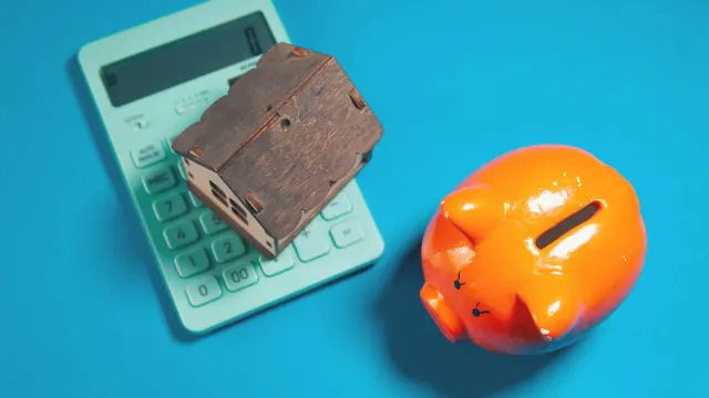 A piggy bank beside a calculator and small house represents money saving challenges.