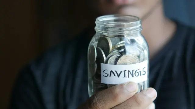 A hand holds a glass savings jar labeled savings for save money jar ideas.