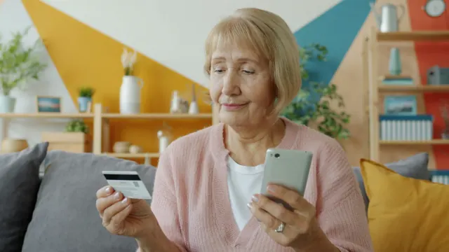 An older woman compares a card and phone, showing why is saving money so hard.